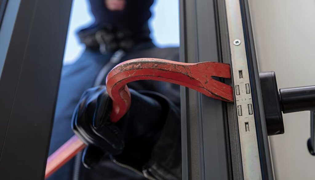 Thief in balaclava holding a crowbar to break a glass window.
